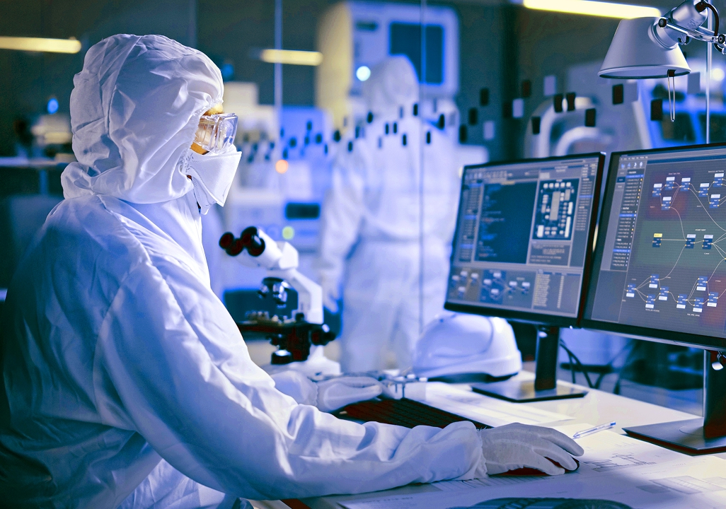 Scientist in cleanroom PPE analyzing data on dual monitors at a lab workstation.