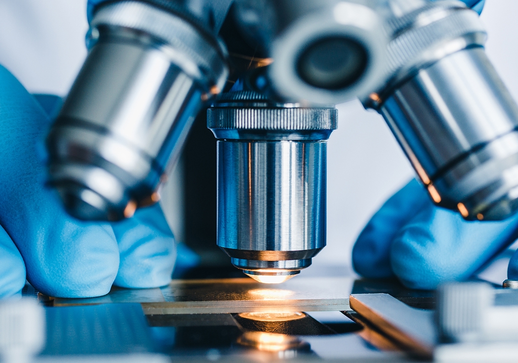 Macro shot of microscope objective lenses focusing on an illuminated slide, with gloved hands adjusting.