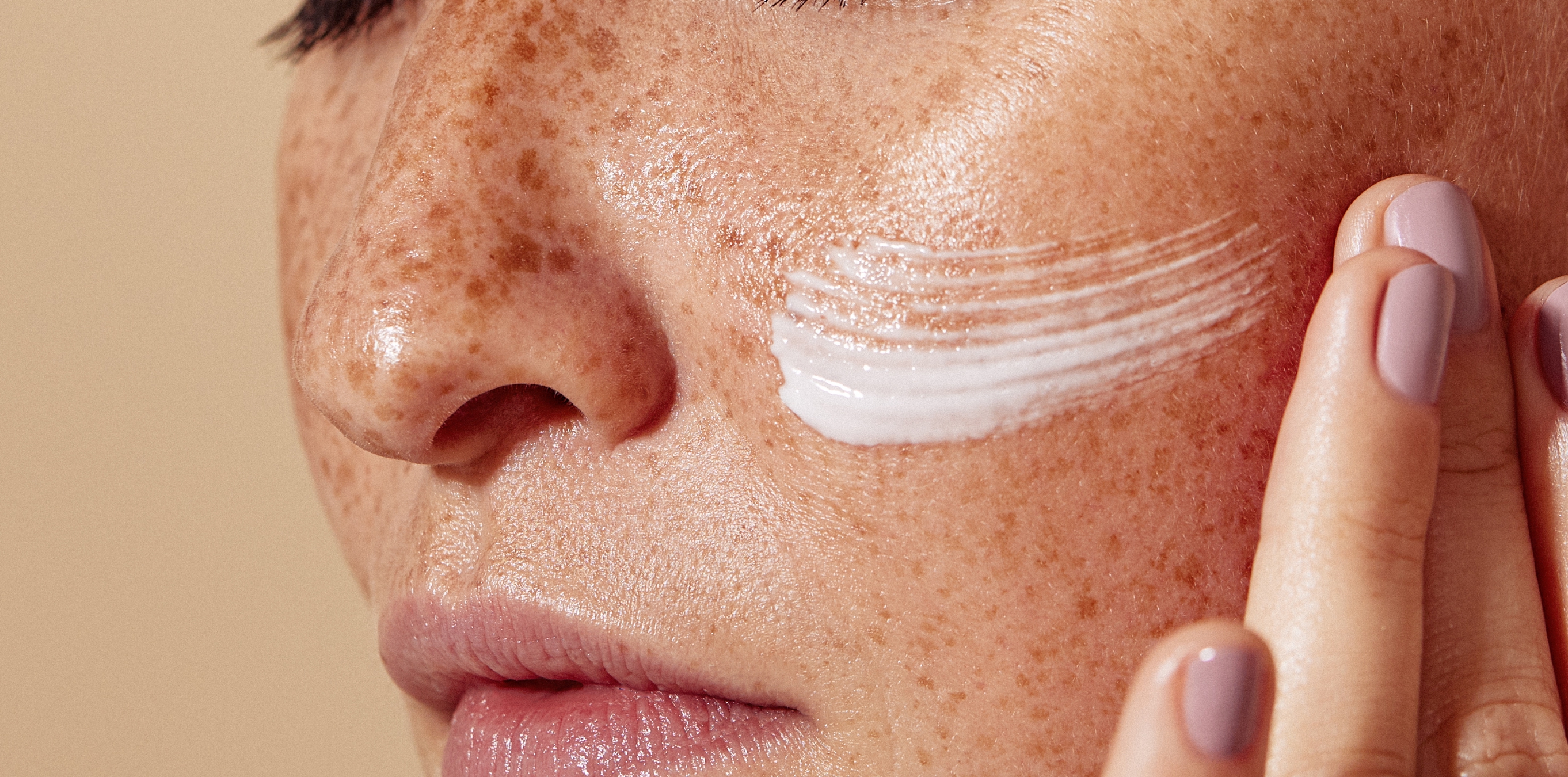 Close-up of a freckled cheek with a swipe of face cream, fingers gently applying it.