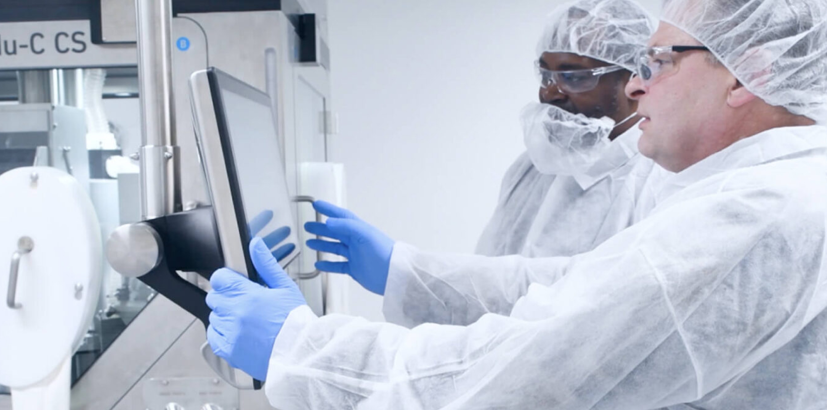 Technicians in cleanroom PPE using a touchscreen control panel inside a pharmaceutical manufacturing suite.