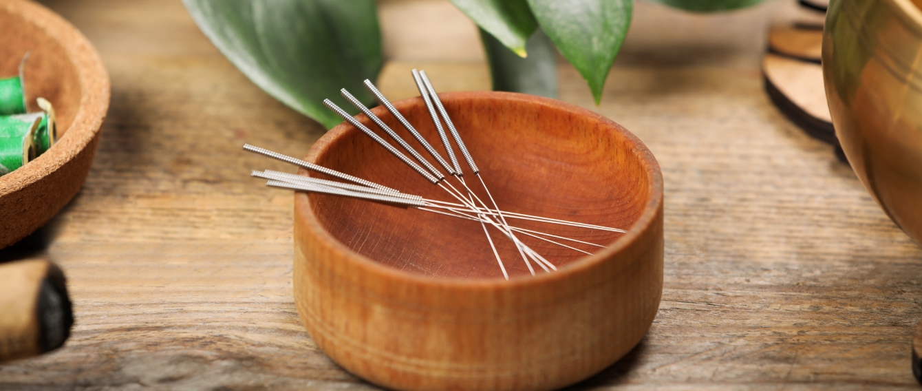 Acupuncture needles resting in a small wooden bowl on a rustic table, with leafy greenery in the background.