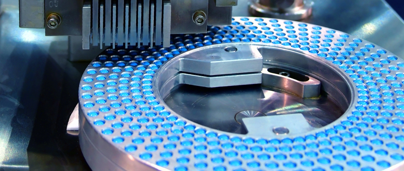 Close-up of a pharmaceutical rotary filling/press machine, showing a circular tray packed with blue tablets or pellets under metal tooling.