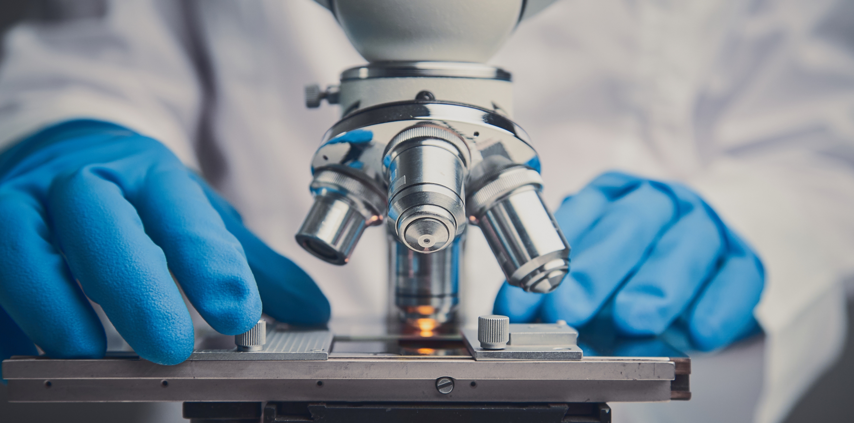 Close-up of gloved hands adjusting a microscope stage and objective lenses.
