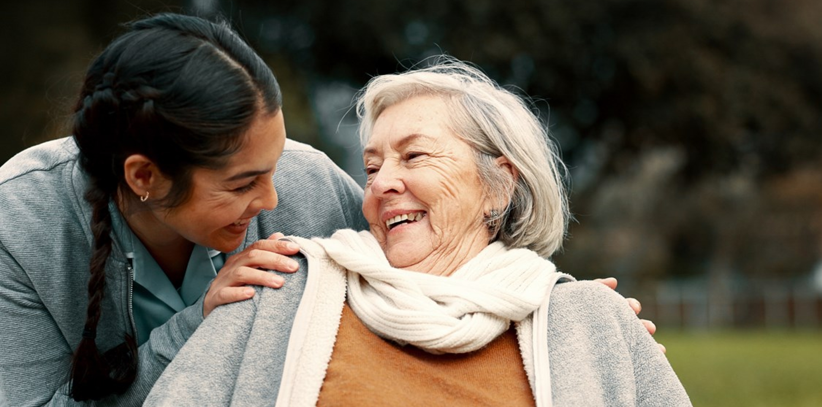 Caregiver smiling with an older woman outdoors, hand on her shoulder.