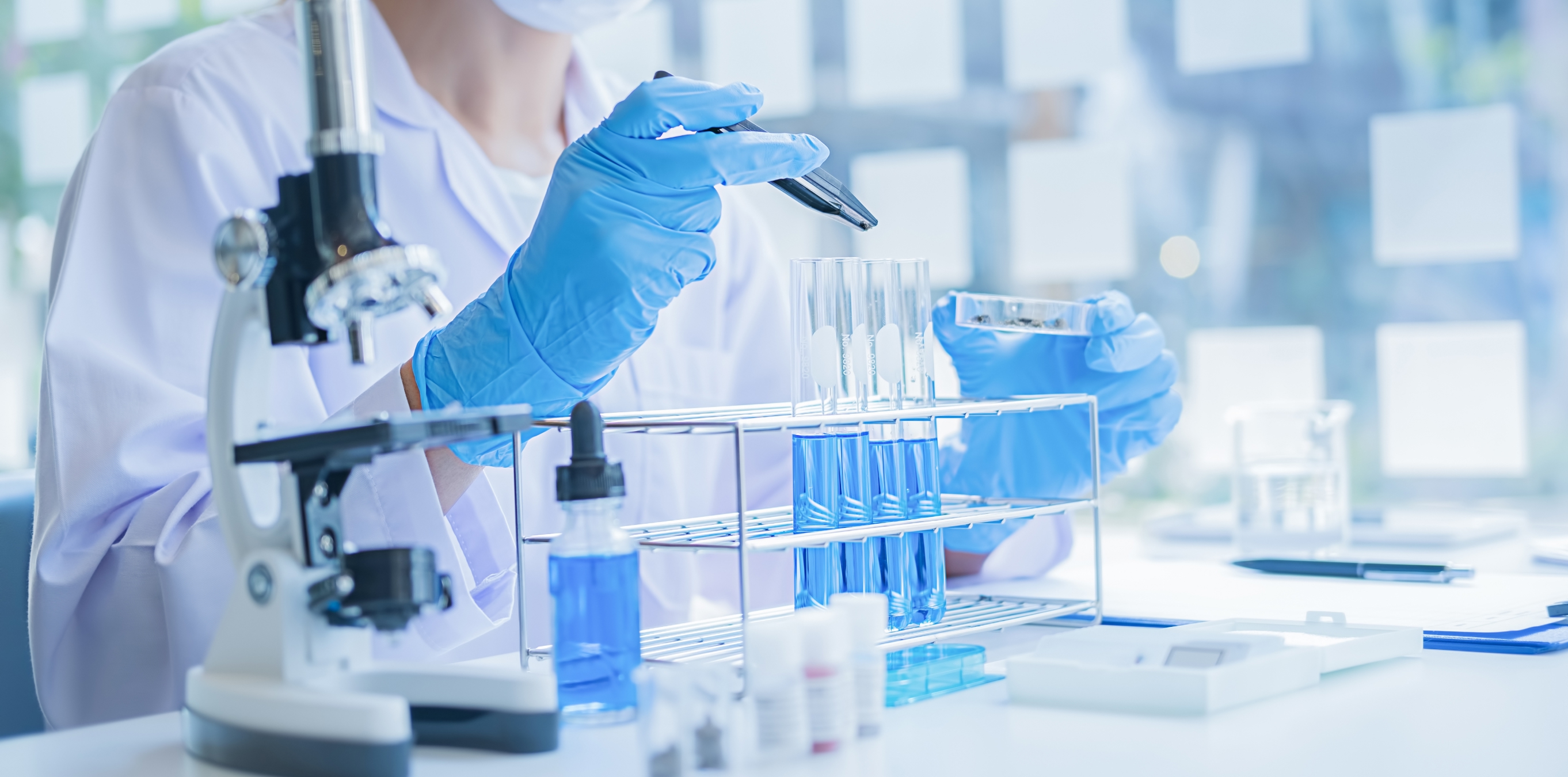 Scientist in gloves handling test tubes with blue samples beside a microscope.