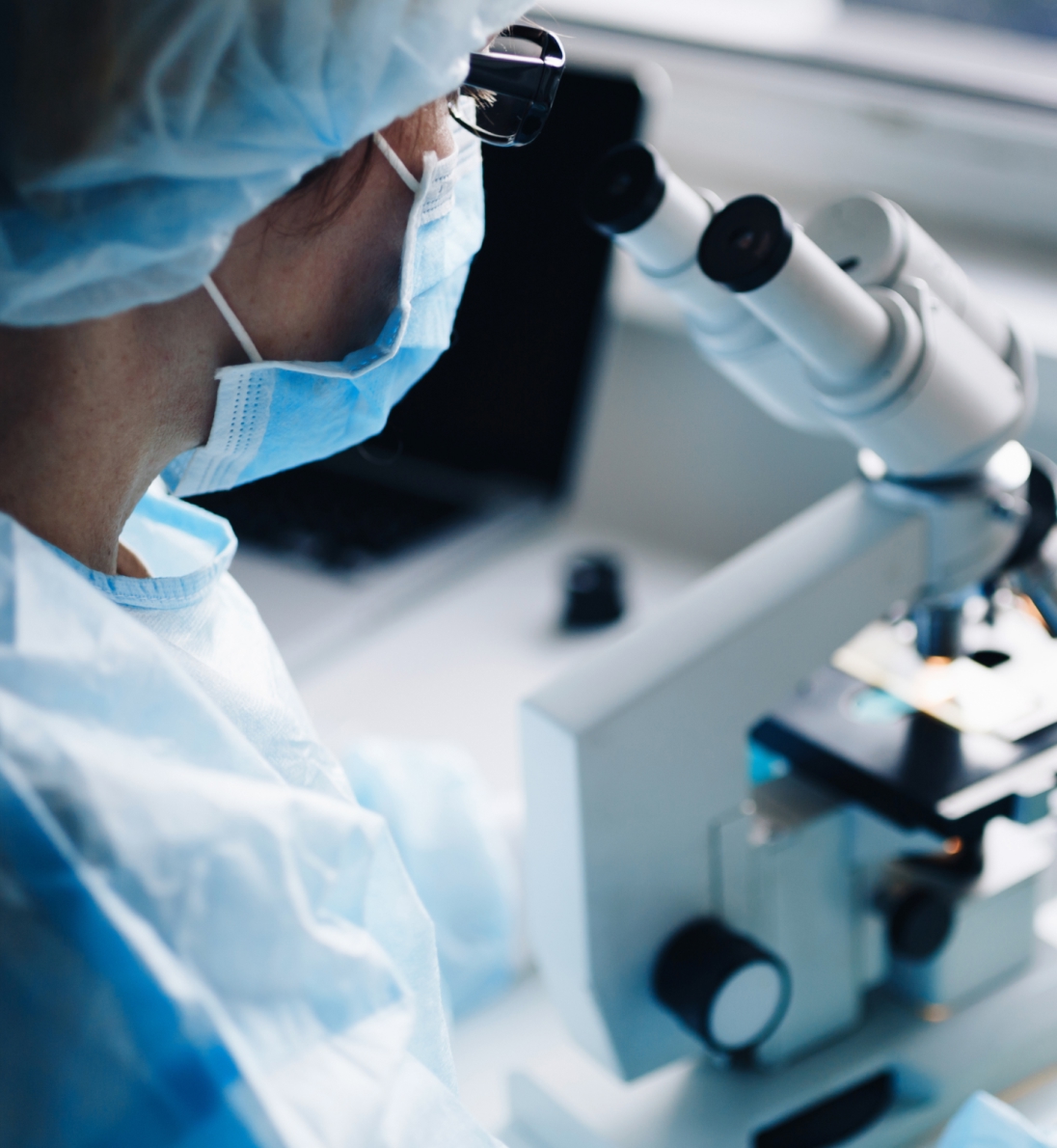 Scientist in mask and gown using a binocular microscope at a lab bench, with a laptop blurred in the background.