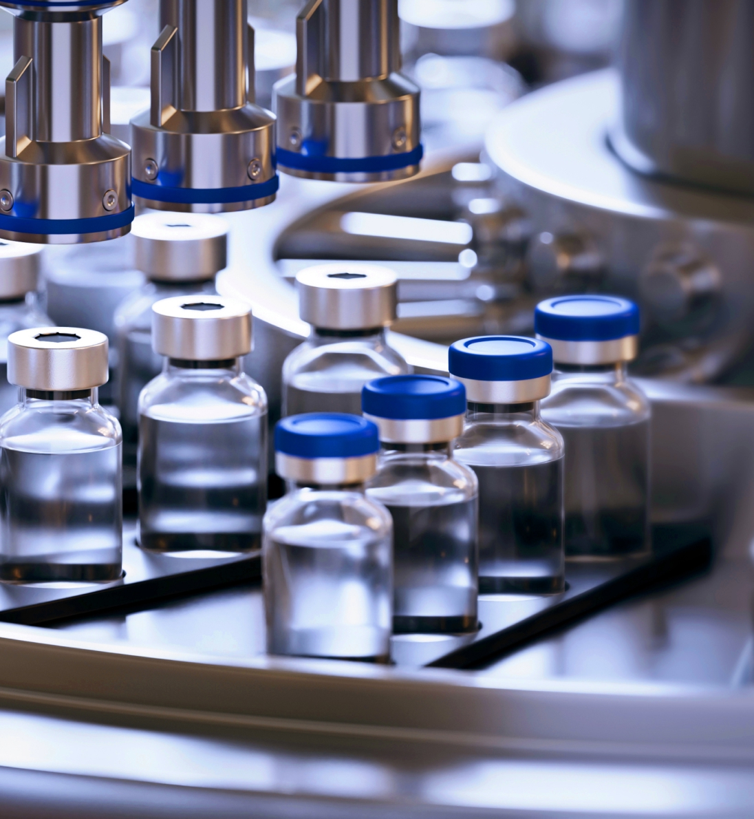 Rows of glass vaccine vials with blue caps move along an automated filling line under stainless-steel nozzles in a sterile pharma facility.
