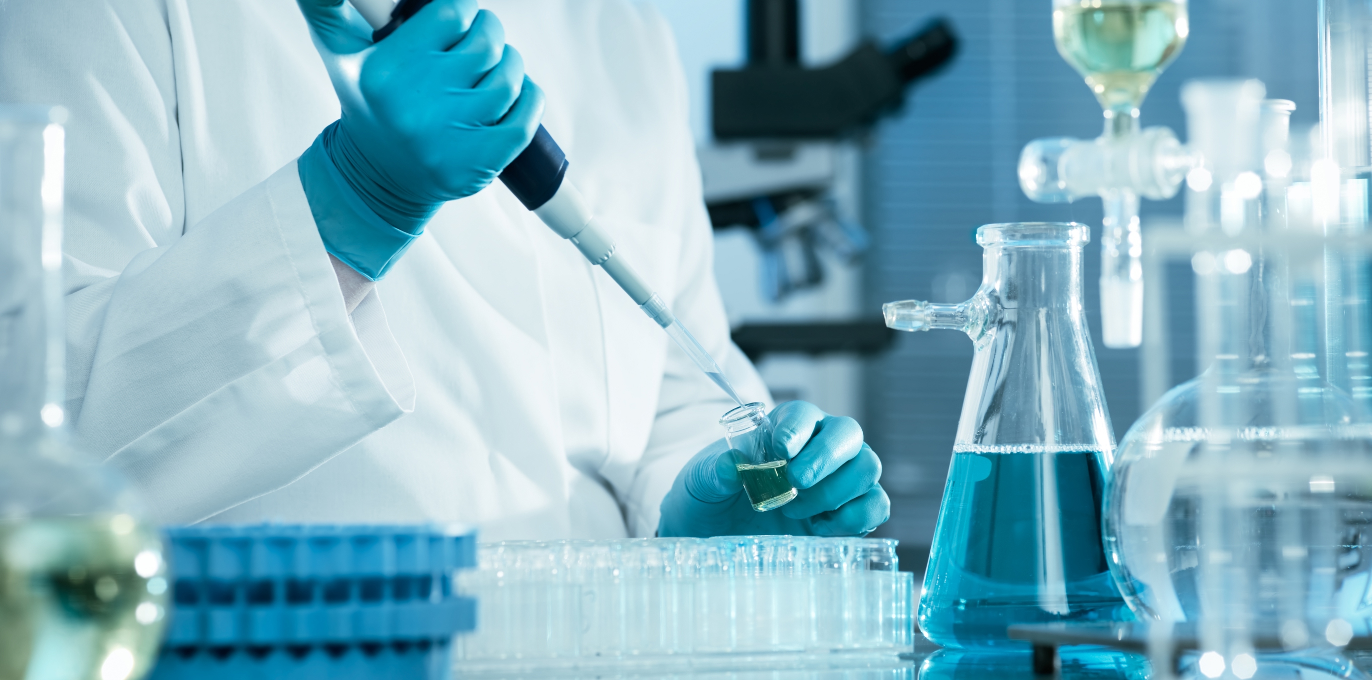 Scientist in lab coat using a pipette to transfer liquid into a vial, with flasks and test tubes on the bench.
