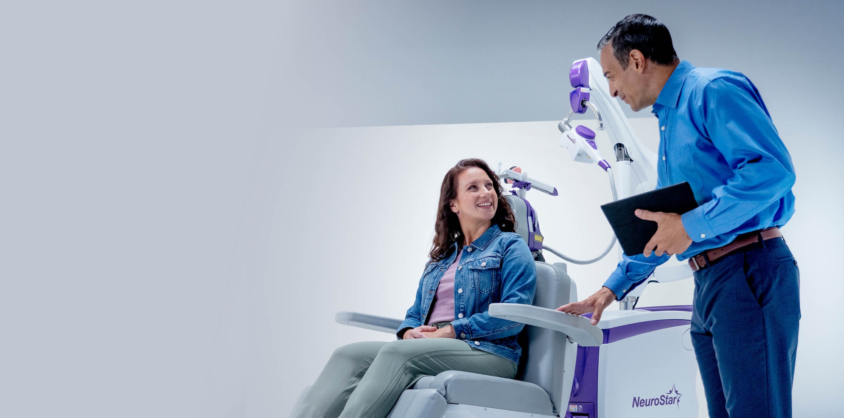 Patient seated in a NeuroStar TMS chair speaking with a clinician holding a tablet.
