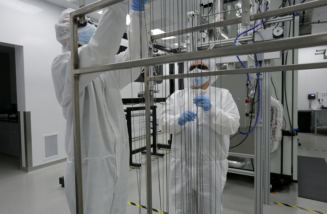 Technicians in cleanroom suits handling thin filaments on a frame inside a sterile manufacturing lab.