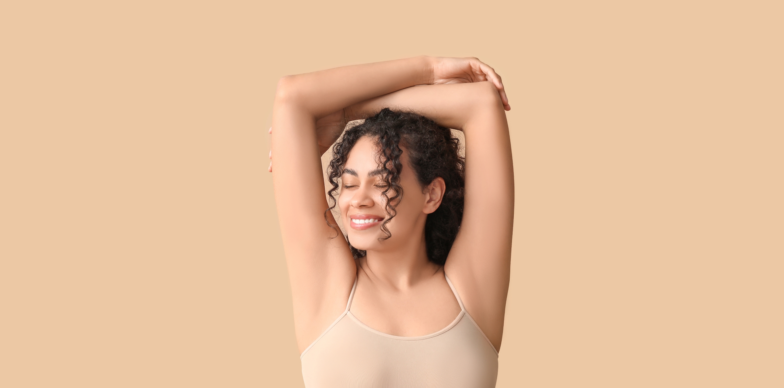 Woman with curly hair smiling and stretching with arms raised against a beige background.