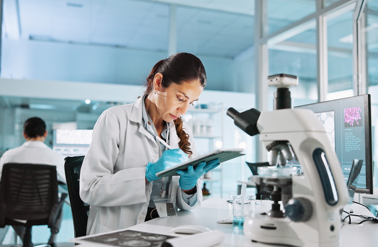 Researcher in a lab coat and gloves recording results on a tablet beside a microscope and monitor.